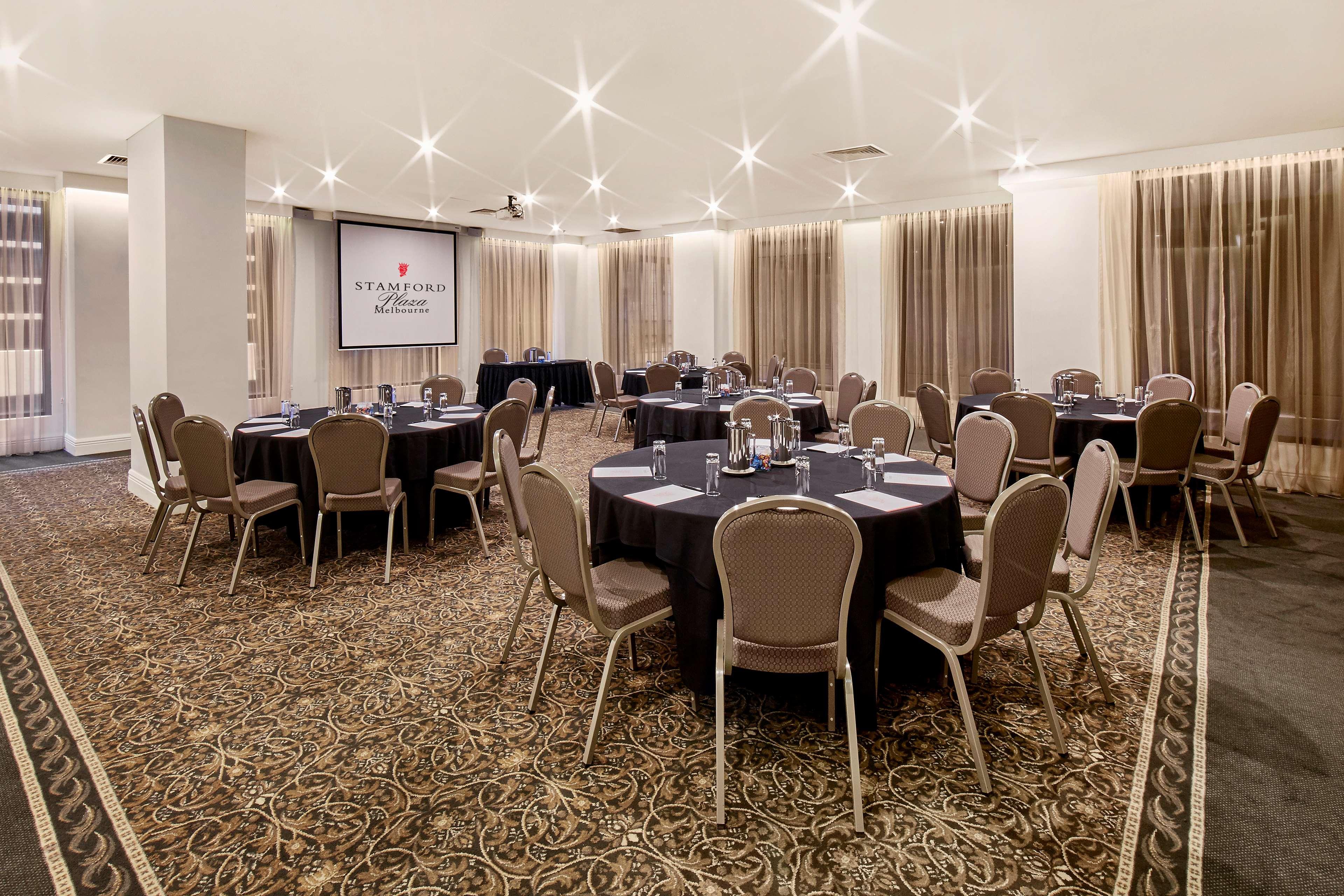 Stamford Plaza Melbourne Hotel Exterior foto The photo shows a well-lit event space set up for a gathering or meeting. There are several round tables covered with black tablecloths, each surrounded by beige chairs. In the background, a large screen displays a logo or branding, likely related to