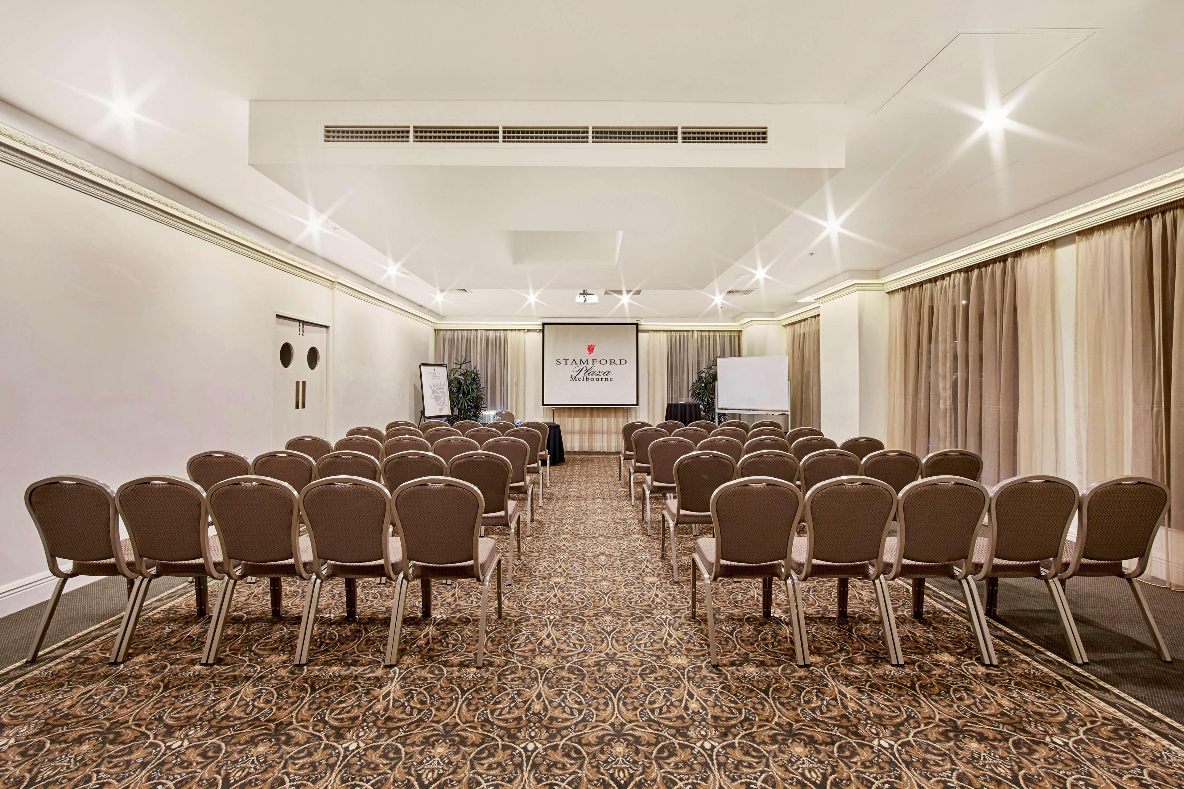 Stamford Plaza Melbourne Hotel Exterior foto The photo shows a conference room set up for an event. It features several rows of beige chairs arranged facing a central presentation area. At the front, there is a screen displaying a logo or title, possibly for a conference or meeting. The walls h