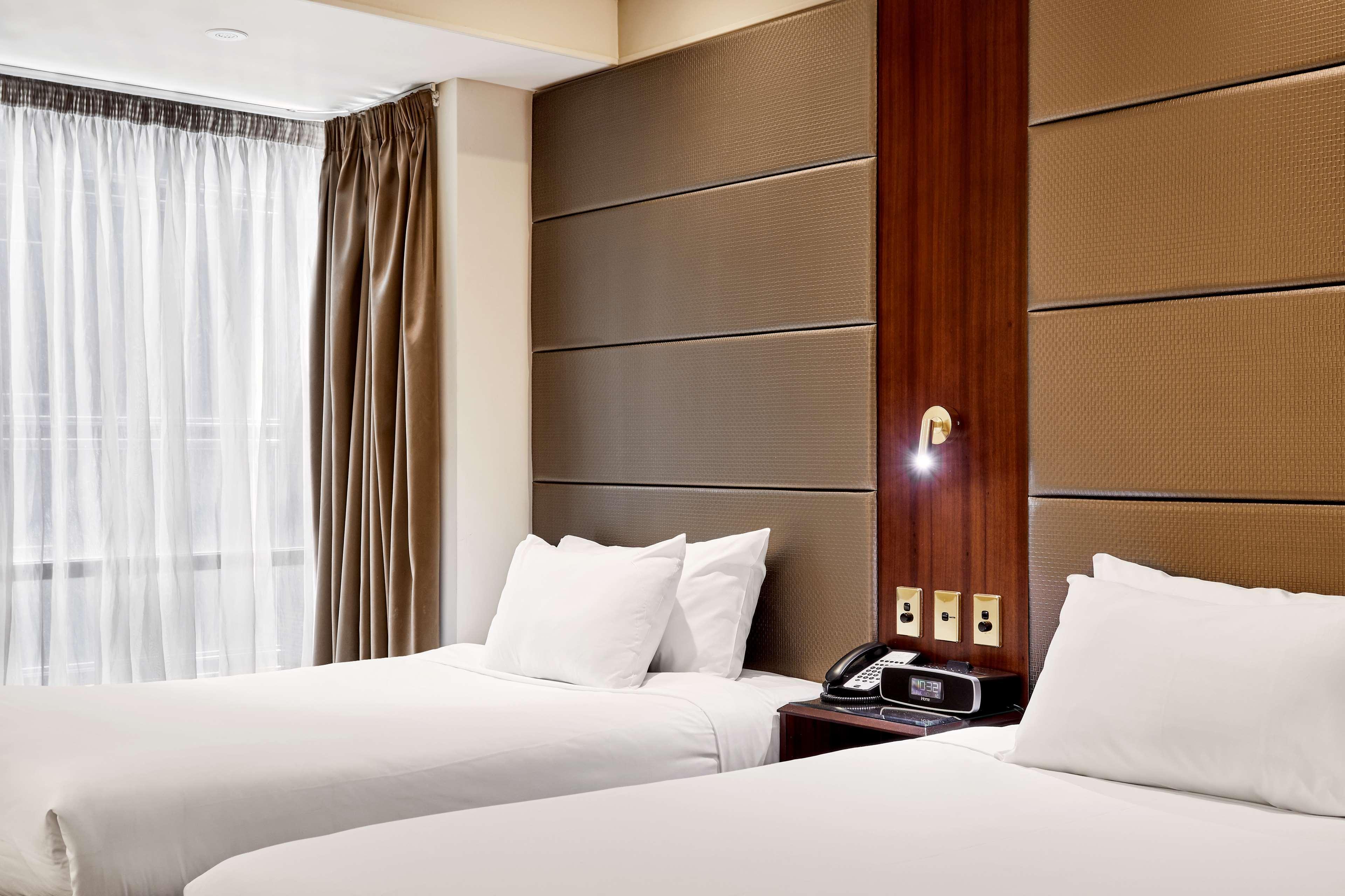 Stamford Plaza Melbourne Hotel Exterior foto The photo shows a modern hotel room featuring two twin beds with white linens and fluffy pillows. The walls behind the beds have a combination of textured panels in neutral colors. There is a bedside table with a lamp and a phone. A large window with