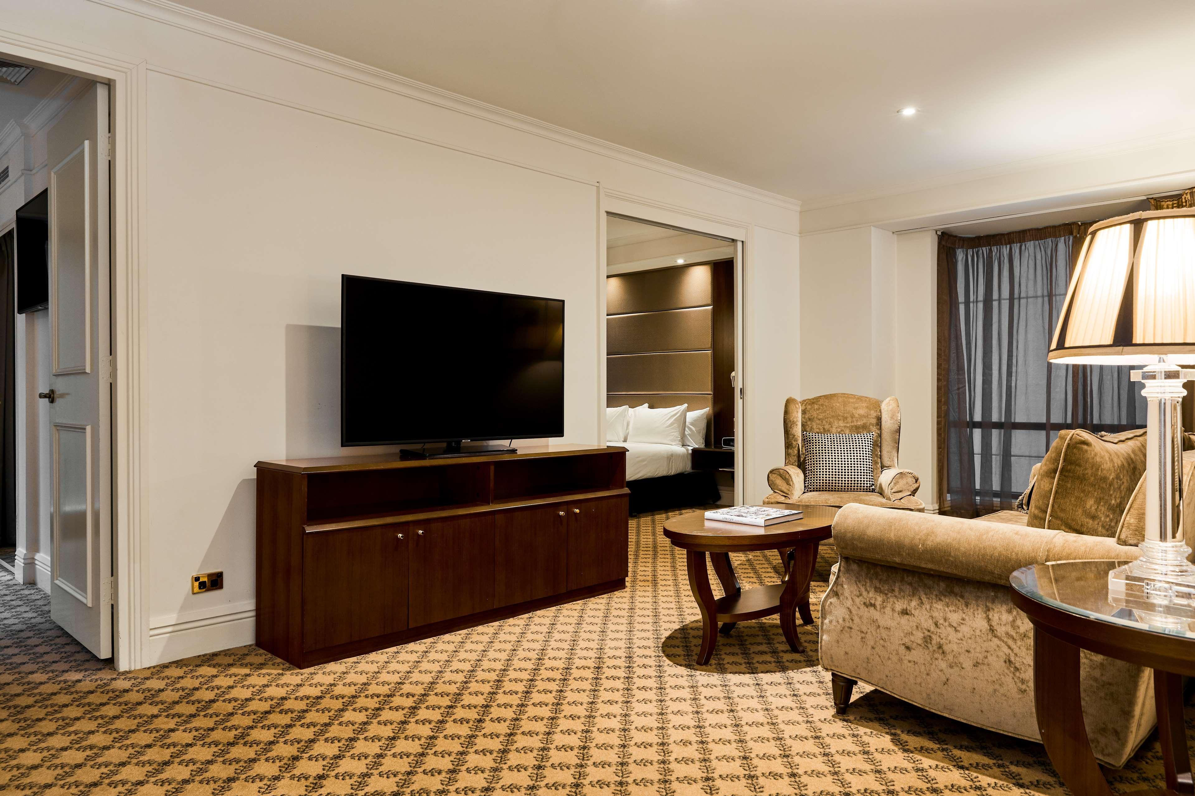 Stamford Plaza Melbourne Hotel Exterior foto The photo shows a well-furnished room, likely a hotel suite or a living area. There is a large flat-screen television mounted on a wooden entertainment unit. In front of the TV, there is a round wooden coffee table. To the side, a comfortable armchai