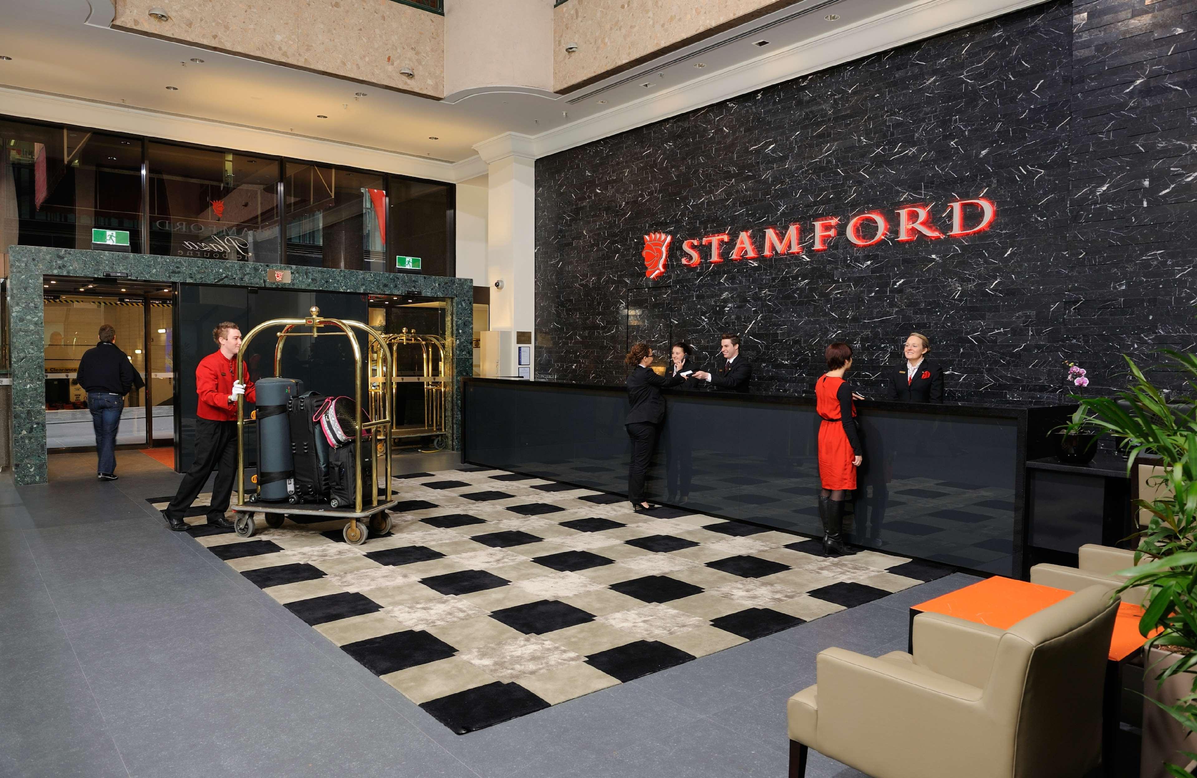 Stamford Plaza Melbourne Hotel Interior foto The photo shows the interior of a hotel lobby, specifically the Stamford Hotel. There is a large reception desk where several staff members are attending to guests. In the foreground, a bell cart loaded with luggage is positioned beside the reception
