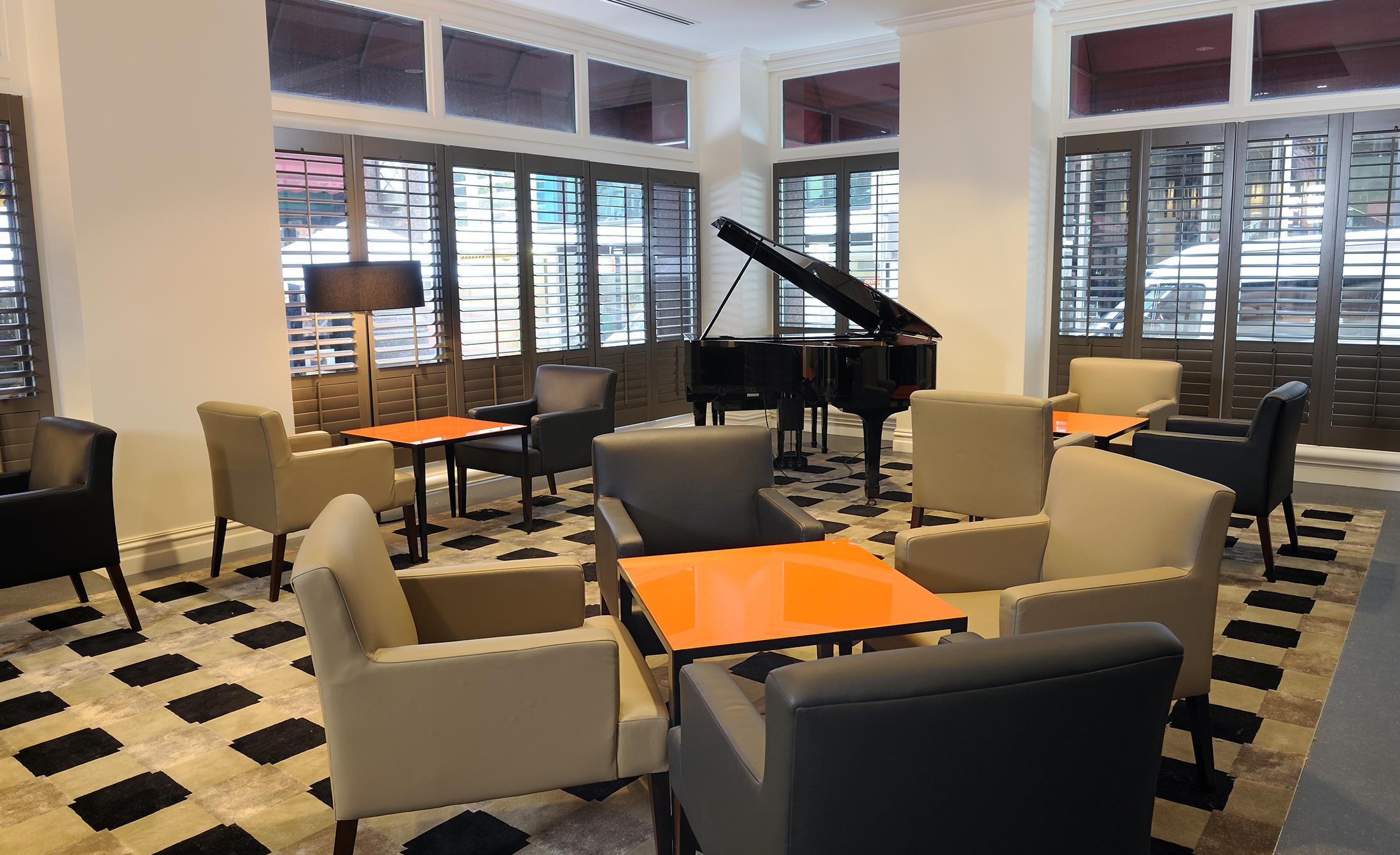 Stamford Plaza Melbourne Hotel Exterior foto The photo shows a stylish indoor lounge area. It features several light gray armchairs arranged around orange square tables. In the corner, there is a black grand piano, adding an elegant touch to the space. The room has large windows with wooden shu