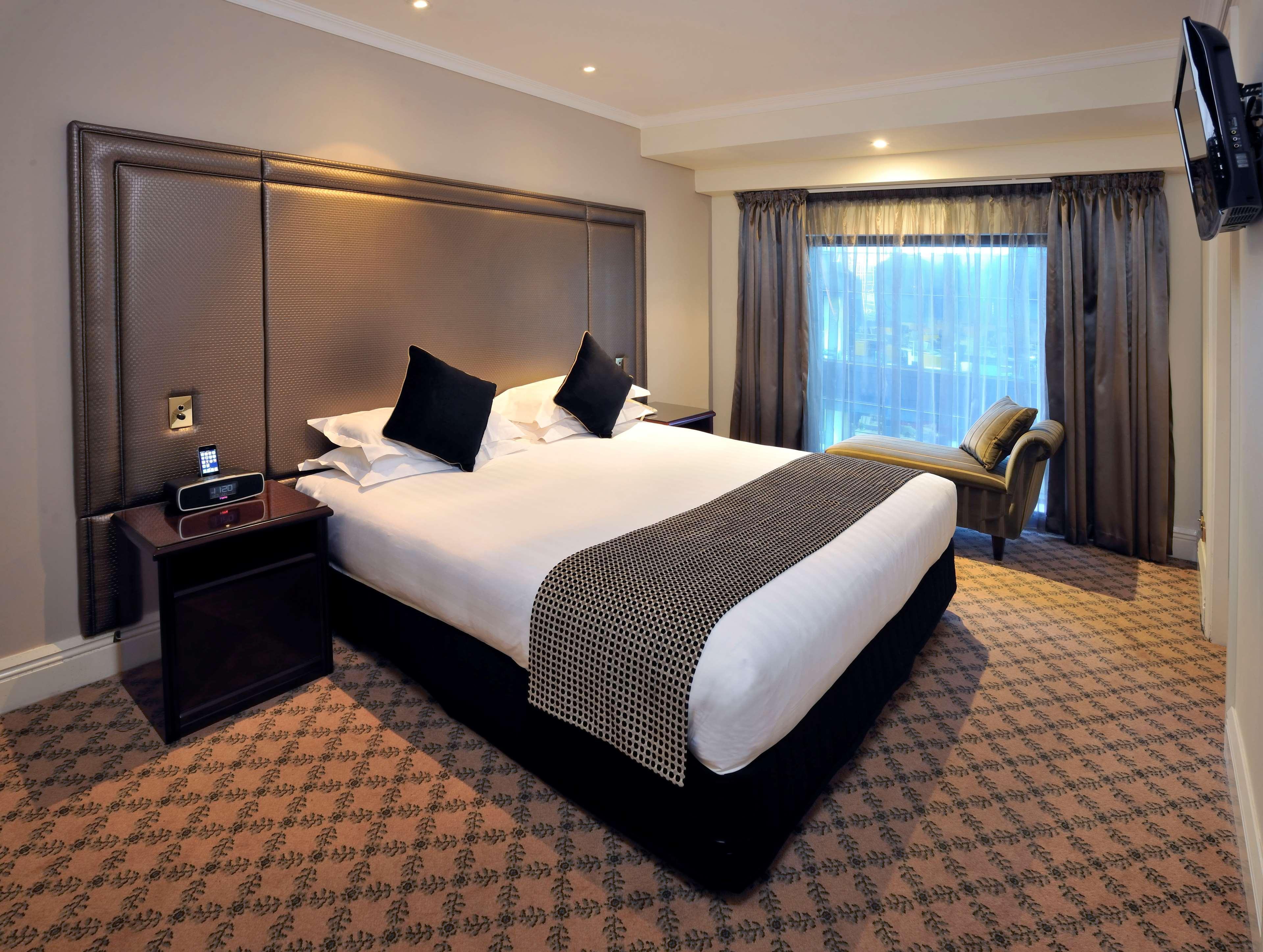 Stamford Plaza Melbourne Hotel Exterior foto The photo shows a hotel room featuring a neatly made king-sized bed with dark pillows. The headboard is upholstered in a dark material, and there is a decorative blanket at the foot of the bed. Beside the bed are two nightstands, each equipped with l