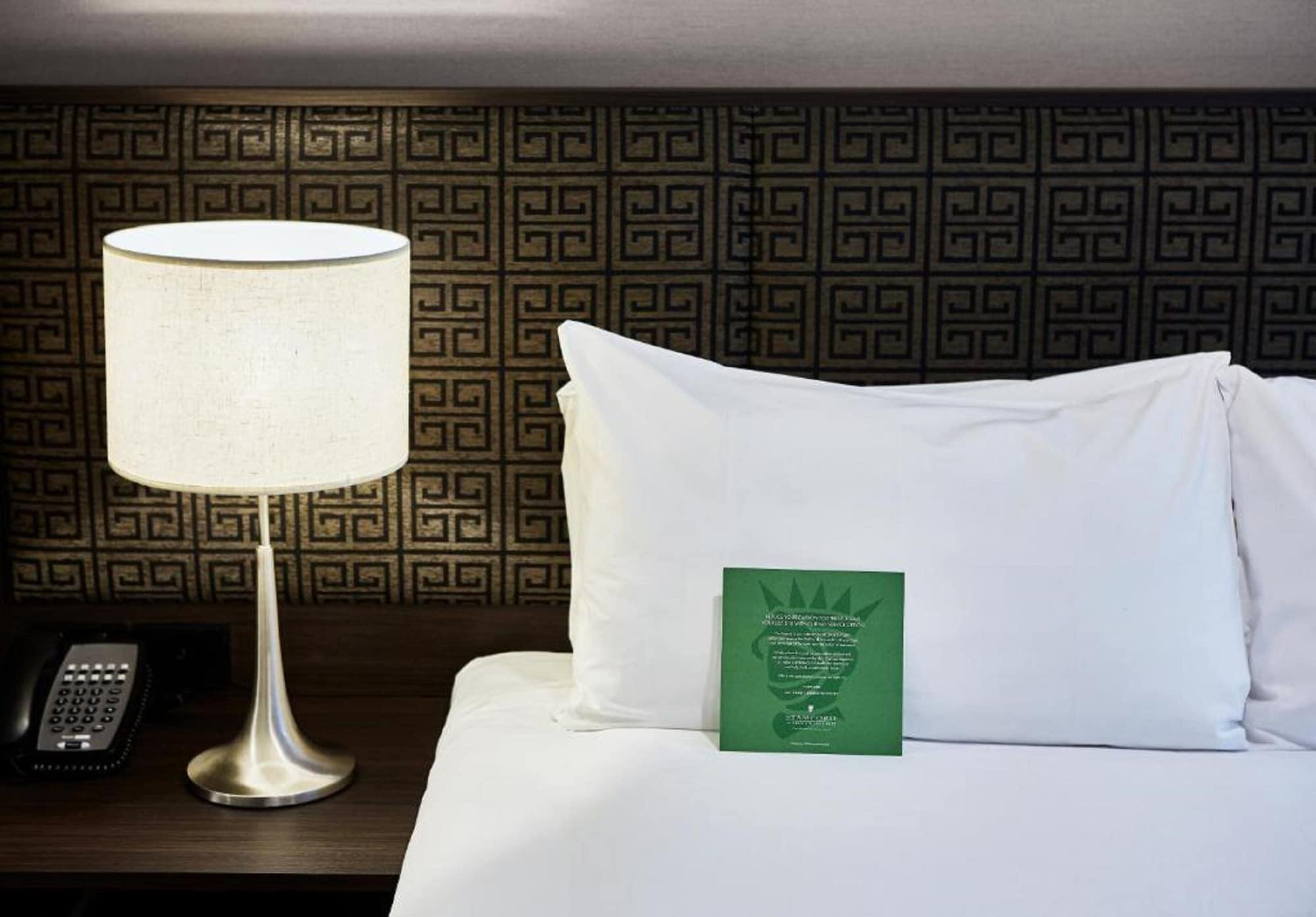 Stamford Plaza Melbourne Hotel Exterior foto The photo shows a neatly made bed with a white pillow and a green item placed on top of it. Next to the bed, there is a stylish table lamp with a lampshade that appears to be white. In the background, you can see a patterned wall, which has a geometr