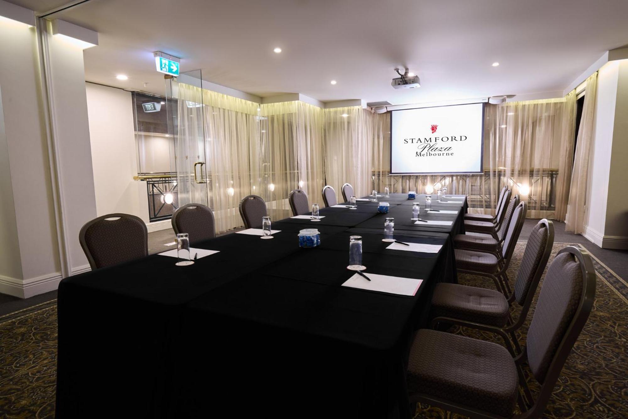 Stamford Plaza Melbourne Hotel Exterior foto The photo shows a well-appointed conference room designed for meetings or presentations. There is a long rectangular table covered with a black tablecloth, surrounded by upholstered chairs. On the table, there are several folded pieces of paper and g