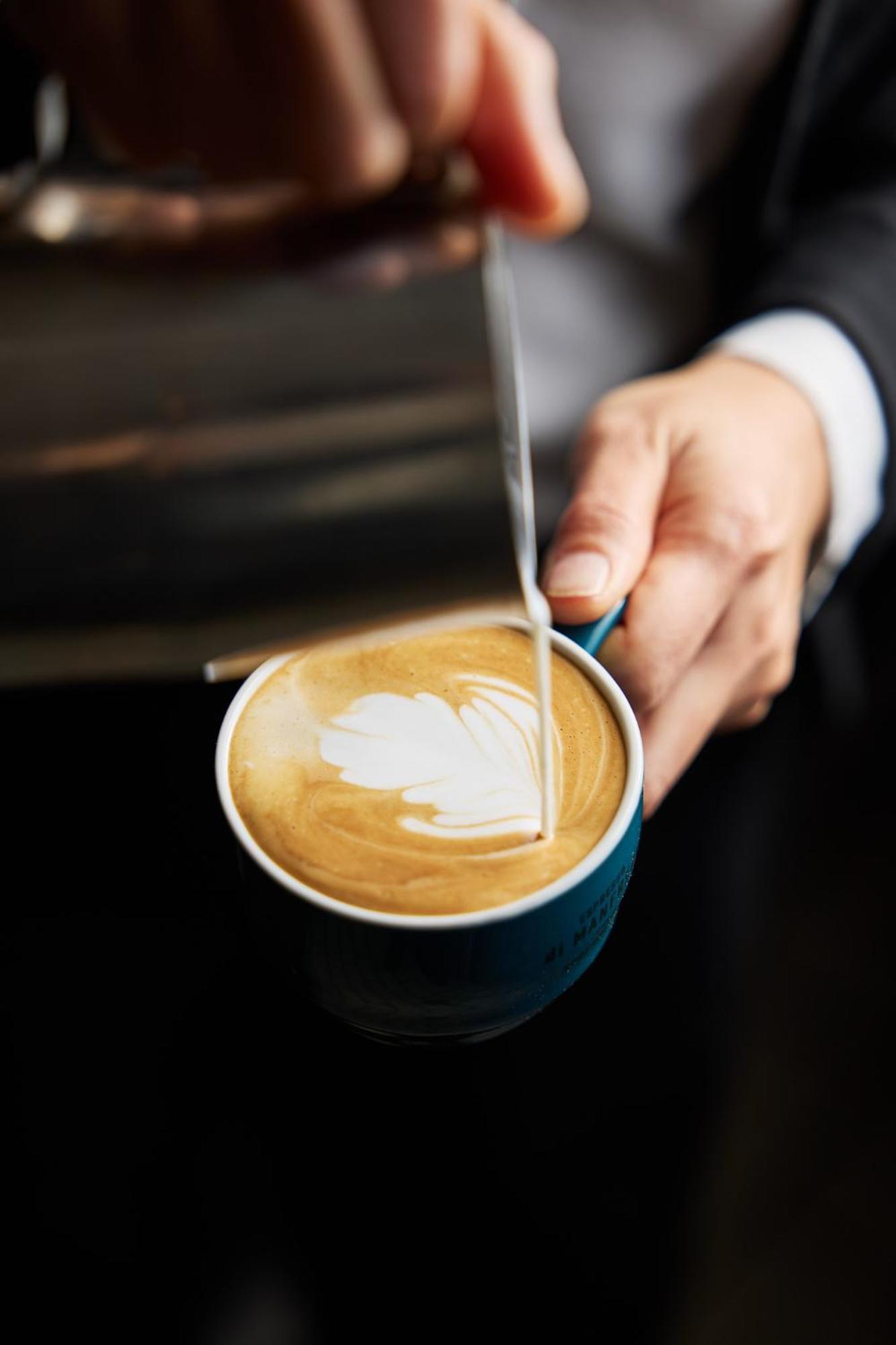 Stamford Plaza Melbourne Hotel Exterior foto The photo shows a close-up of a hand holding a cup of coffee with a latte art design on the surface. The coffee appears to be a creamy, caramel color, likely a latte or cappuccino. A small stream of milk is being poured into the cup, creating a heart