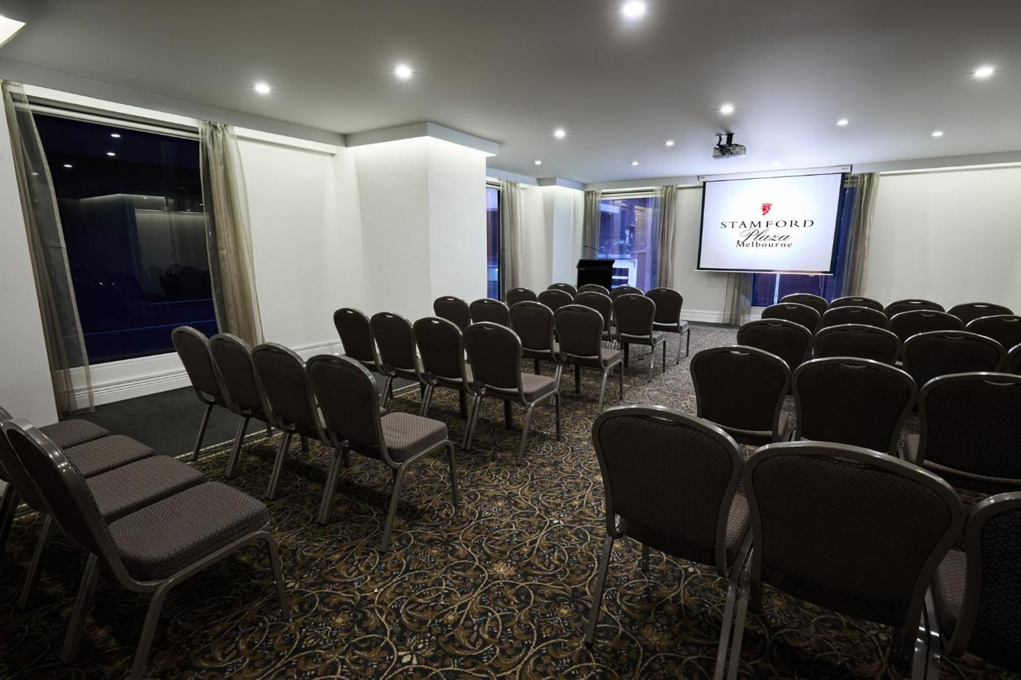 Stamford Plaza Melbourne Hotel Exterior foto The image shows a conference or meeting room set up for an event. There are rows of gray chairs arranged facing a large screen displaying the name "Warford House." The room is well-lit with overhead lighting and has large windows covered with light-c