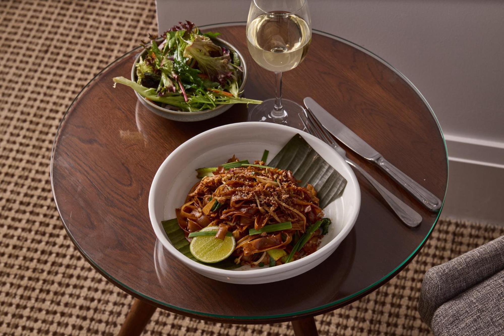 Stamford Plaza Melbourne Hotel Exterior foto The photo shows a meal served on a wooden table. In the foreground, there is a bowl of stir-fried noodles garnished with green onions and a lime wedge, set atop a banana leaf. Next to it, there is a bowl of mixed green salad. A glass of white wine si